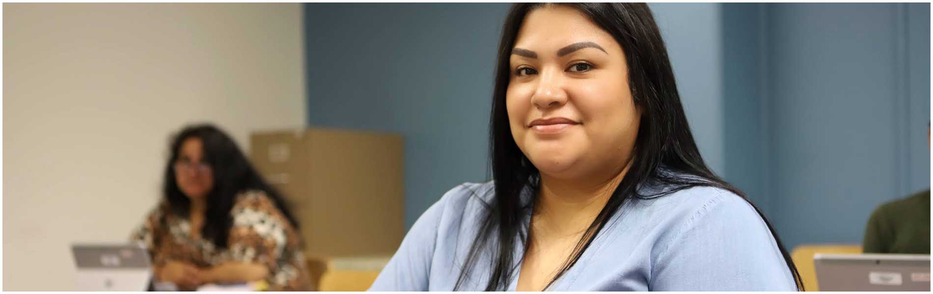 为大学做准备 Student sitting at the table, smiling at the camera with a student in the background looking on