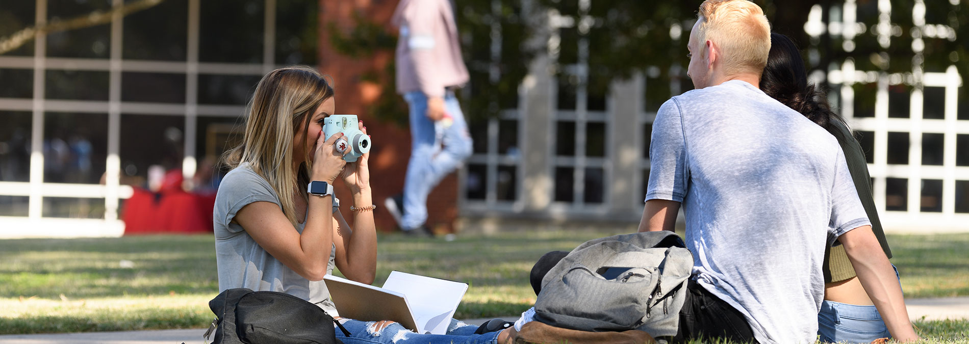 Campus Tour Student sitting on campus grounds, taking a picture of two friends sitting across from the student.
